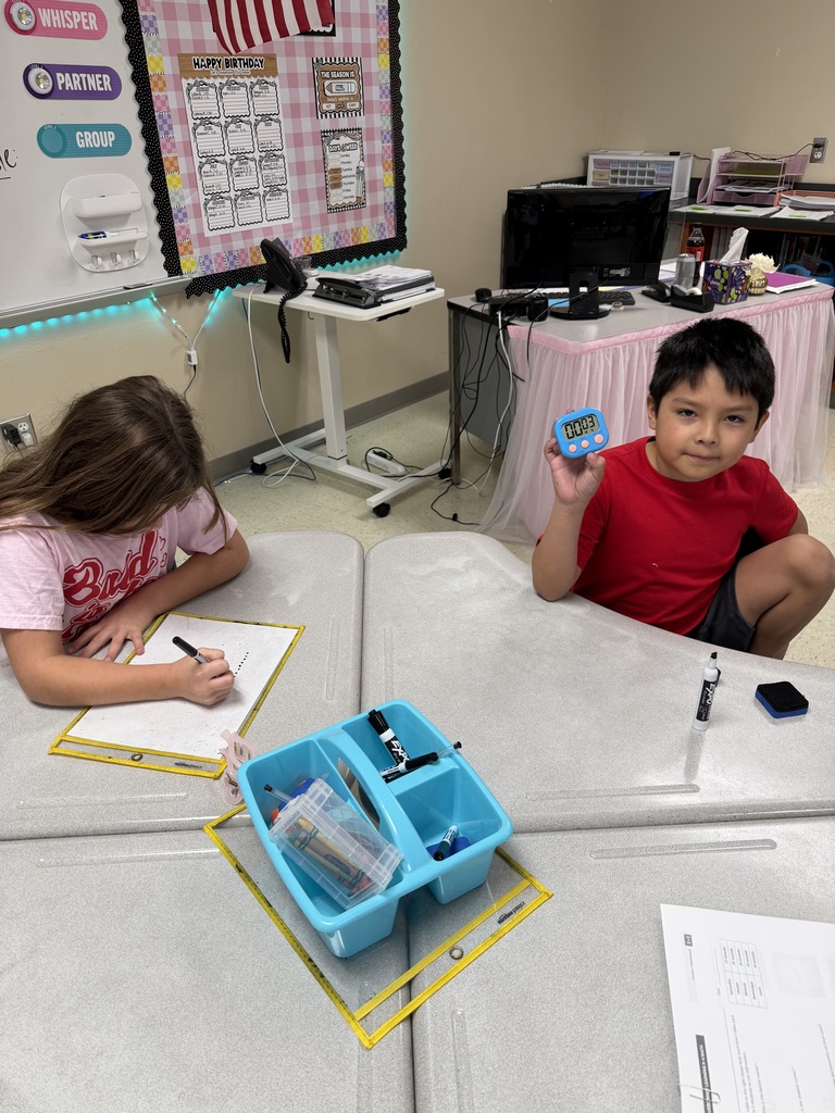 Two children, a boy and a girl, sit at a table in a classroom, each writing on a paper.