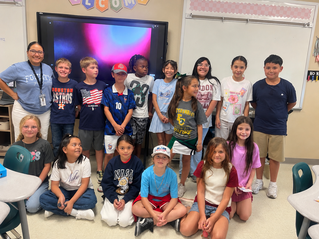 Kids and a woman standing and sitting in a classroom in front of a large screen.