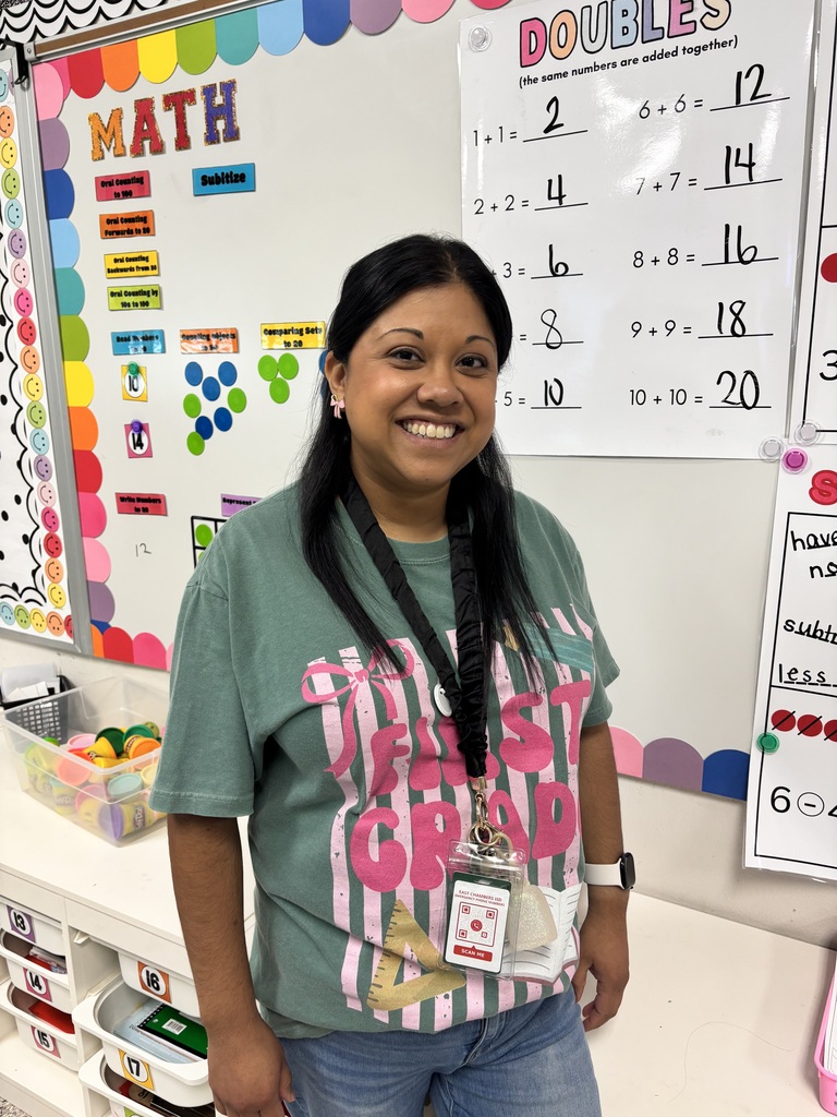 A woman with a smile stands in front of a classroom board. She wears a green t-shirt with white text and a black lanyard.