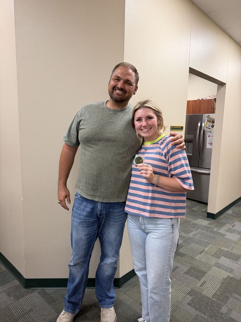 Two people stand in an office hallway, one holding a medal. One wears a green shirt and blue jeans, the other wears a striped shirt and white pants.