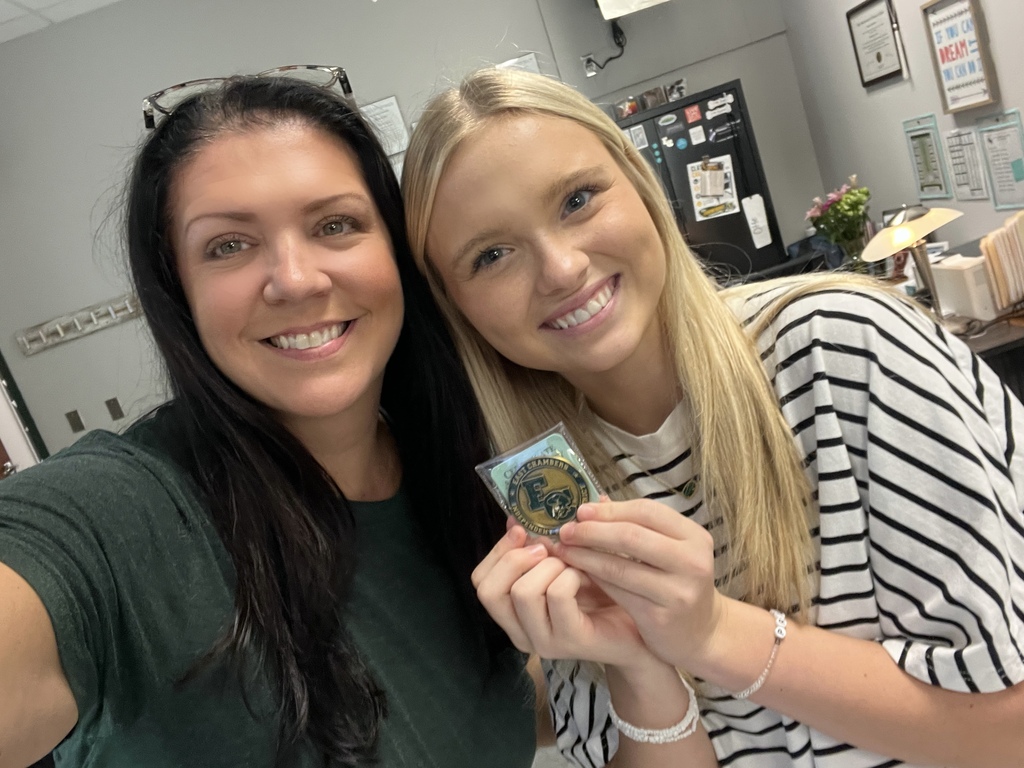 Two women smile in an office. One holds a small, circular item. A refrigerator, framed pictures, and a plant are in the background.