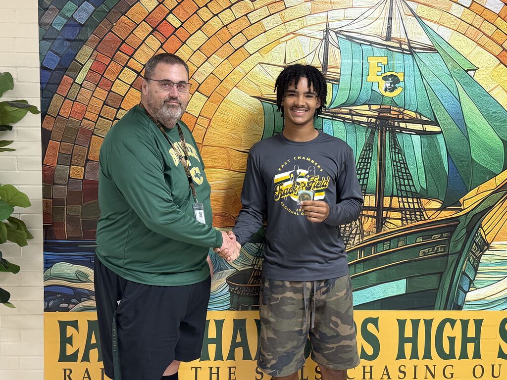 Two individuals stand, one giving a thumbs up, in front of a mural depicting a ship and text.