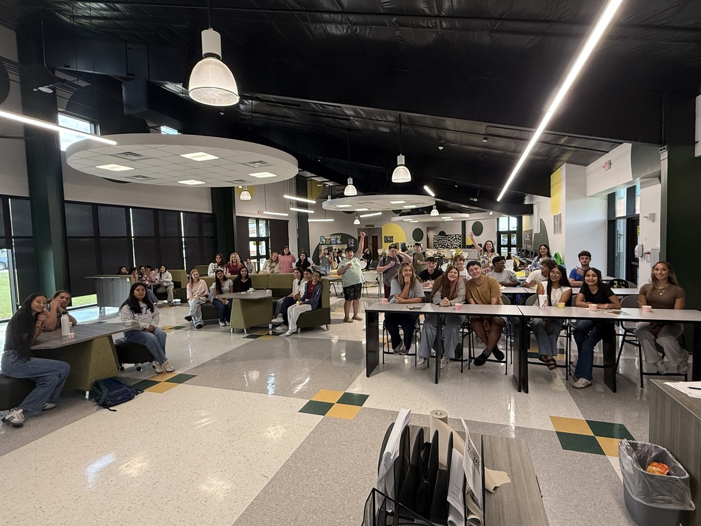 Many people seated at tables in a cafeteria, under fluorescent lights and large ceiling circles.