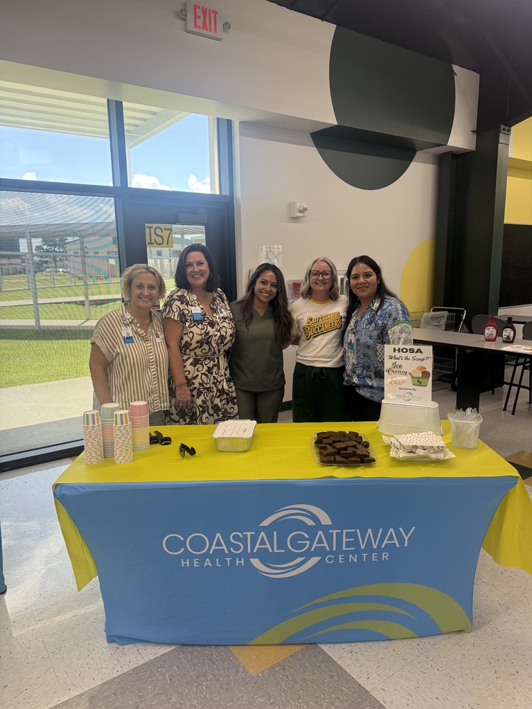 Five women stand behind a table with Coastal Gateway Health Center logo. There are food items on the table. A grassy field is visible outside the door.