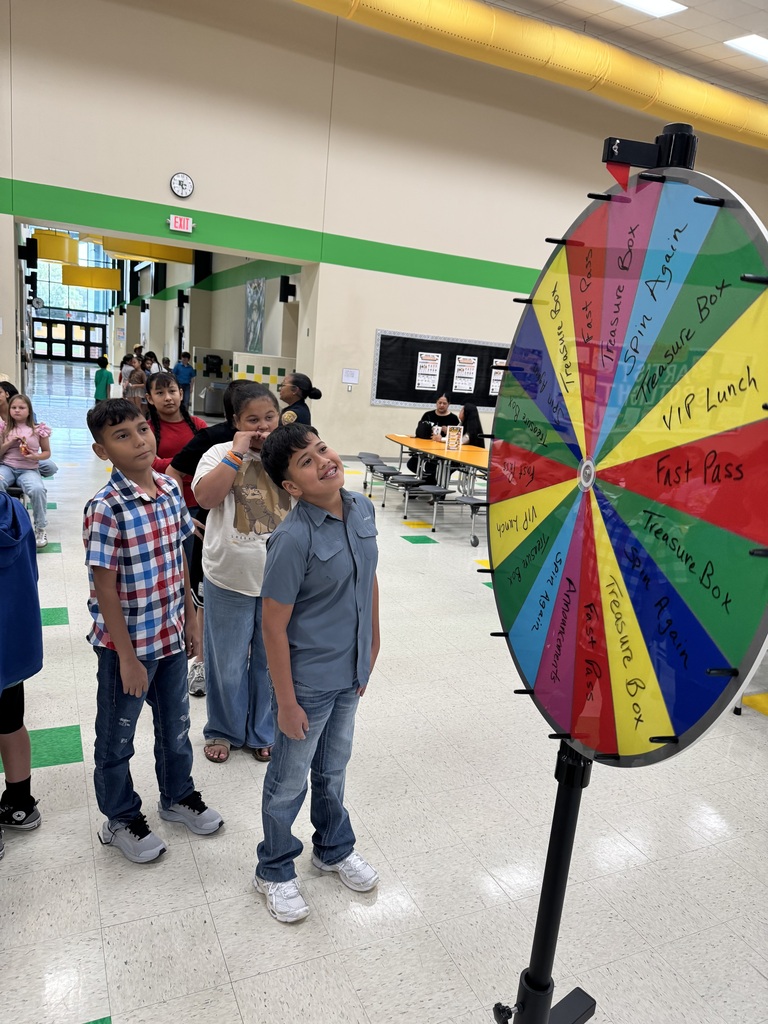 A group of children in a hallway around a large colorful wheel. One child points at the wheel.
