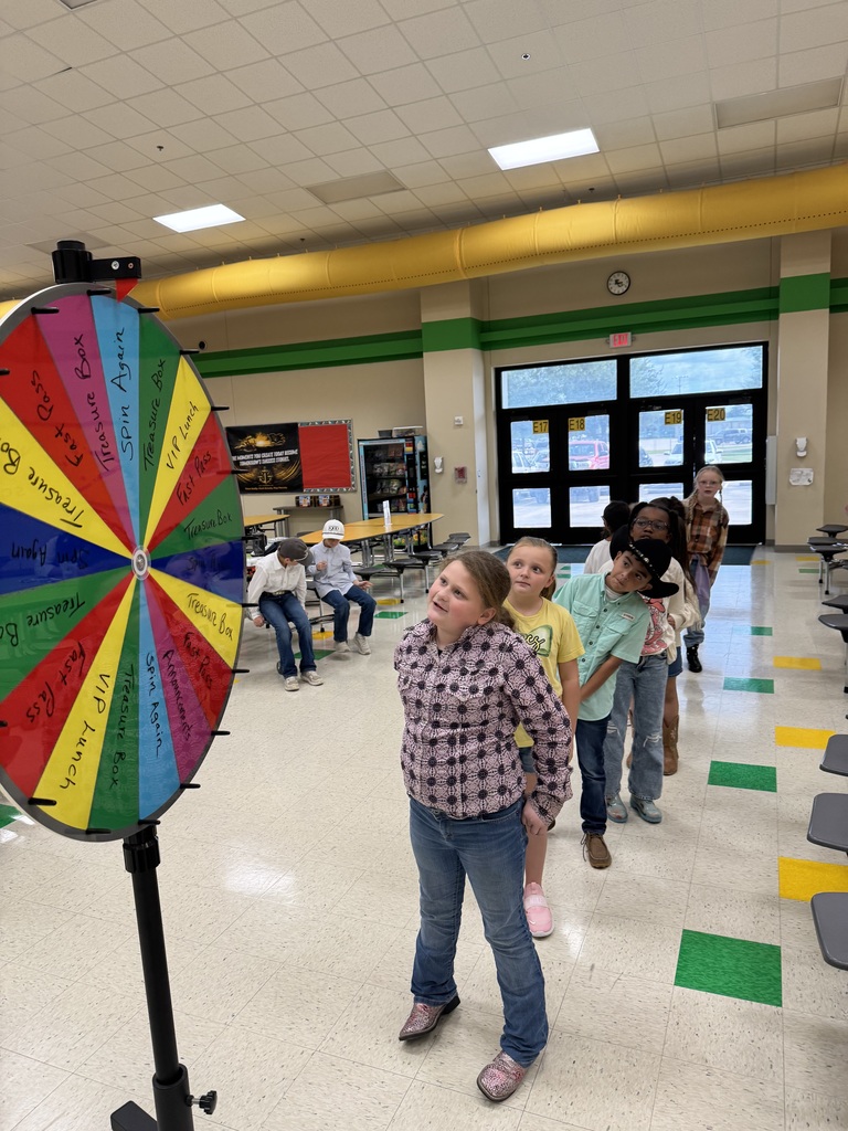 A group of children with a woman stand near a colorful wheel inside a school hallway.