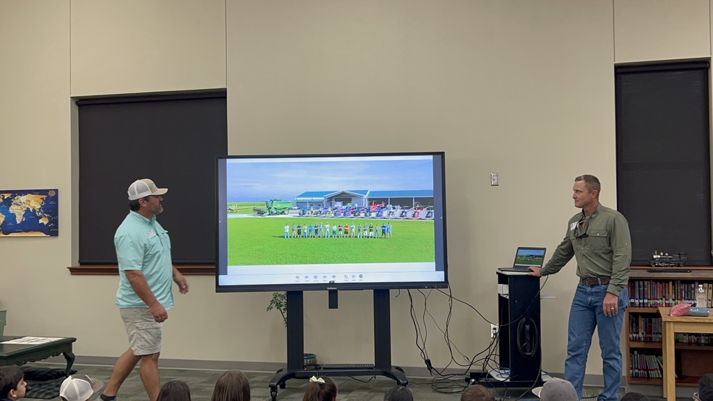 Two men in front of a large screen displaying a field and audience. One man wears a hat, and the other holds a laptop.