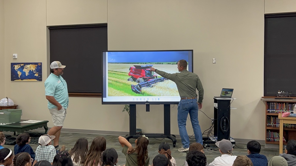 Two people, one pointing at a large screen, stand in front of a classroom full of students.
