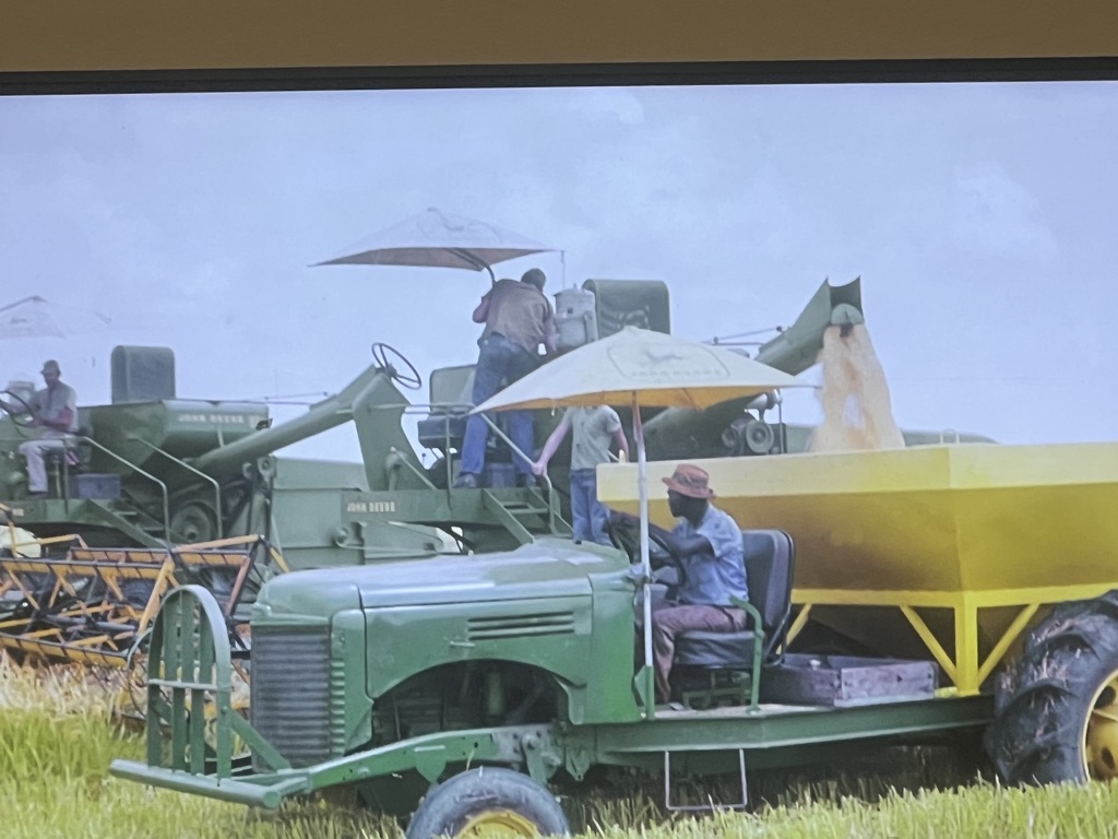 Agricultural machinery in a field with a man in a hat driving a tractor, another man standing on a harvester.