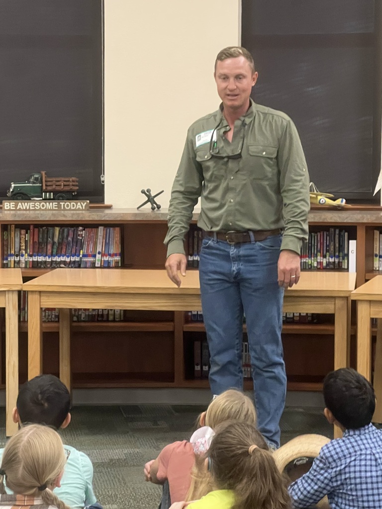 A man stands in front of children. He wears a green shirt and blue jeans. Behind him is a desk with books and a blackboard.