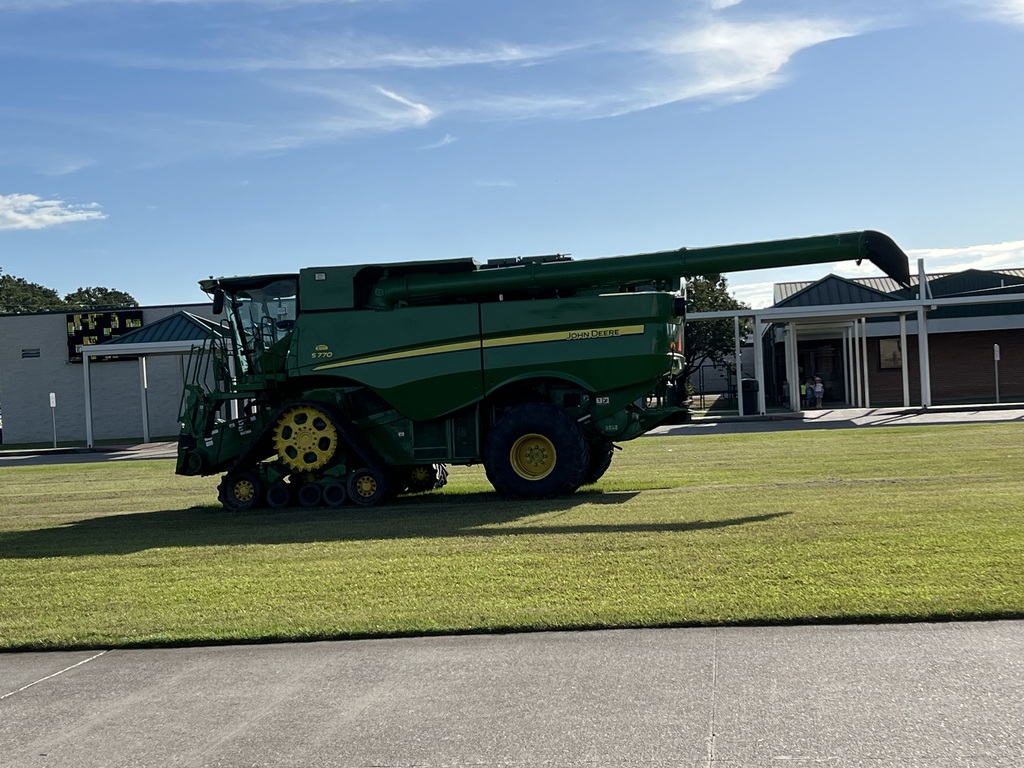 A green harvester is parked on a lawn in front of a building under a partly cloudy sky.