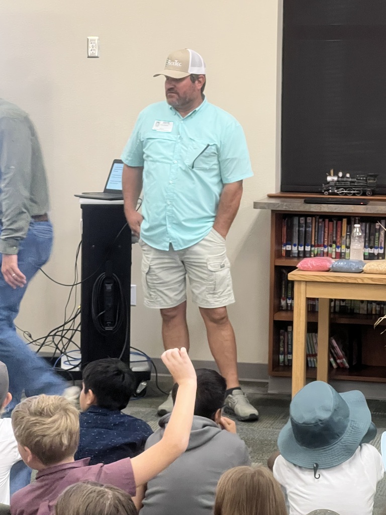 Man standing with hands in pockets, speaking to seated children in a classroom with a bookshelf.