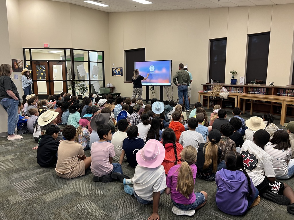A group of students sits on the floor in a room. A man stands before a screen. Several windows are on the wall.