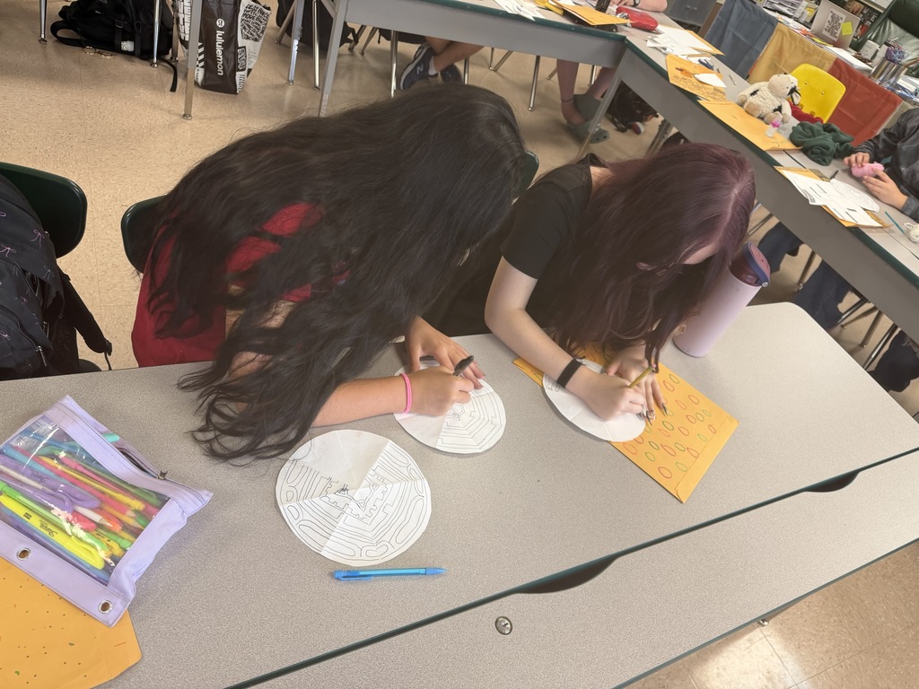 Two students at a table with supplies, one using a marker on a round paper, the other working on a circular paper.