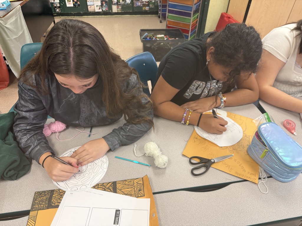 Three people sit at a table with paper crafts, scissors, yarn, and a lunchbox. One is drawing while the other writes.