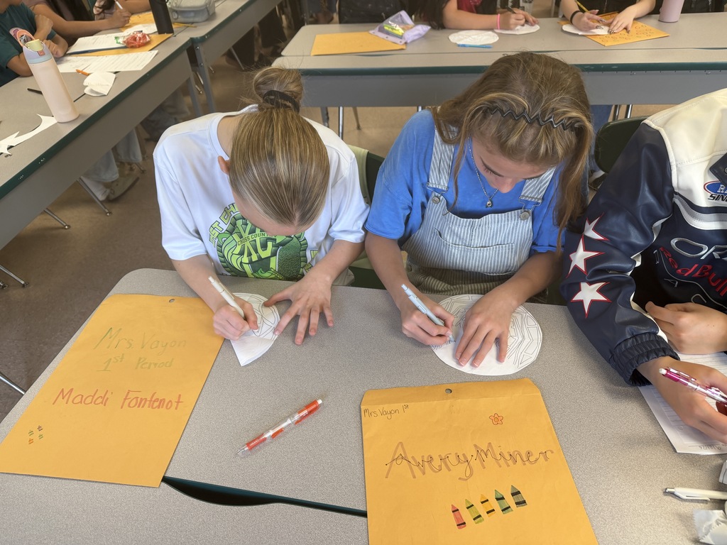 Three students in a classroom, writing on papers. One has "Avery Miner" on the paper.