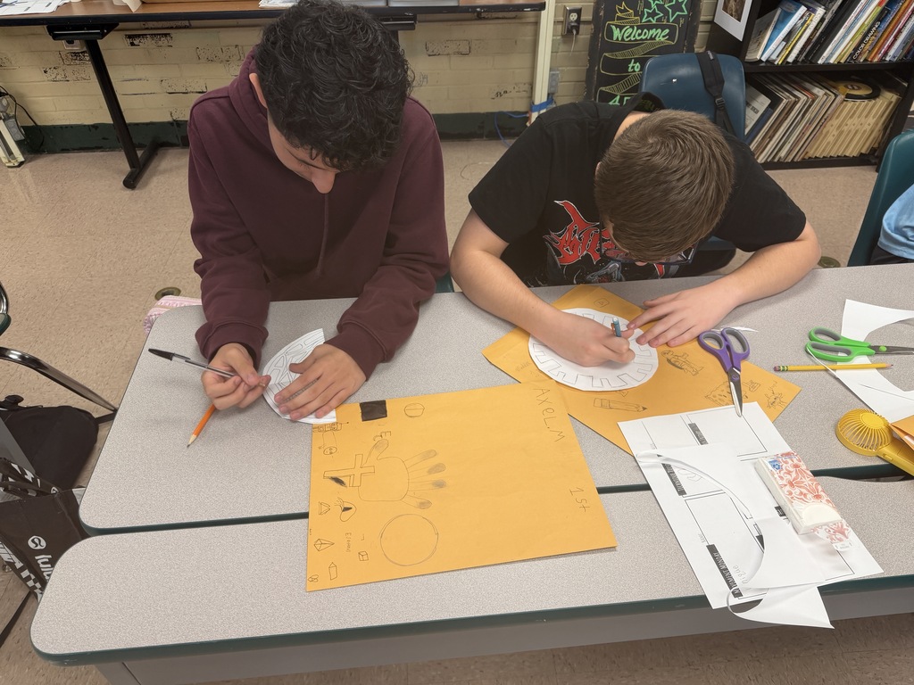 Two students work on a craft project using scissors and paper on a table. The table is filled with craft supplies.