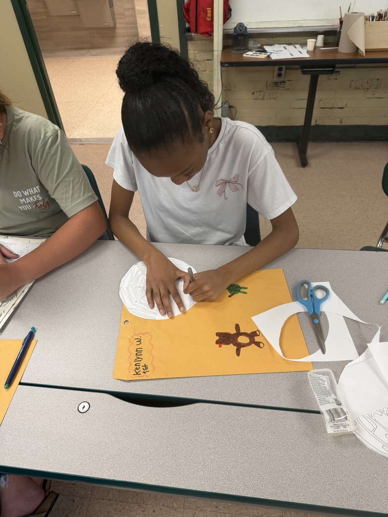 A person sits at a desk, working on a paper with scissors and a pencil. Another person sits nearby.