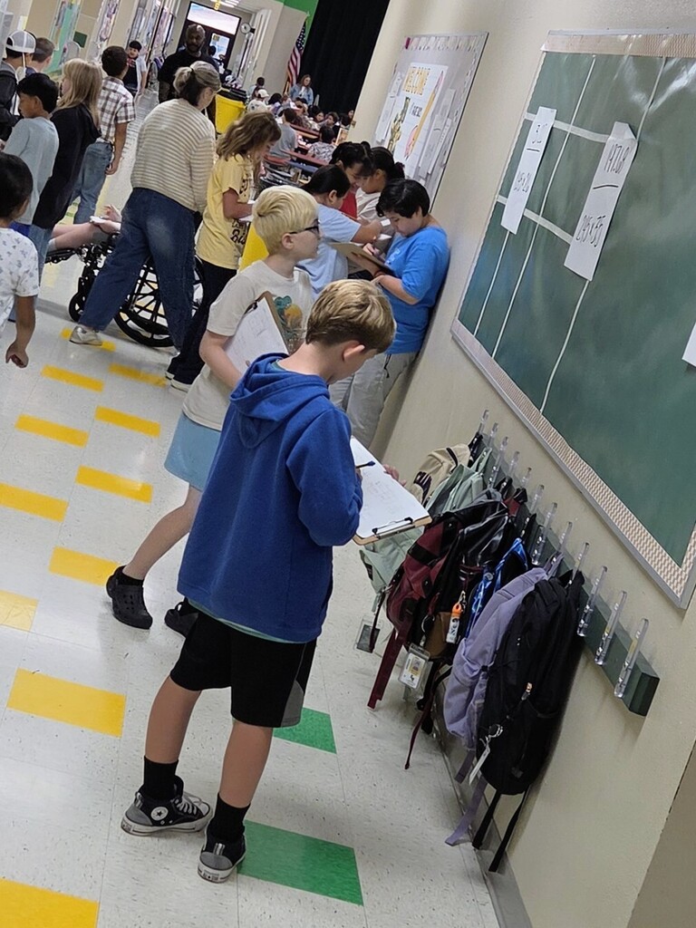 A hallway with yellow and green floor markings. A boy in a blue hoodie leans over a desk. A green board and clothes on hooks are on the wall.