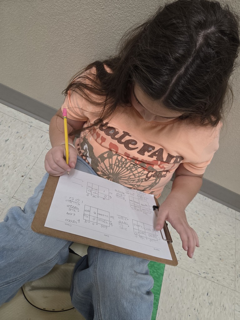 Child seated, holding a pencil, writing on a paper placed on a clipboard. Background: tiled floor.