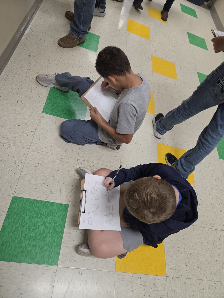 Two students seated on the floor, each writing on a piece of paper. One has a gray shirt, the other a blue one.