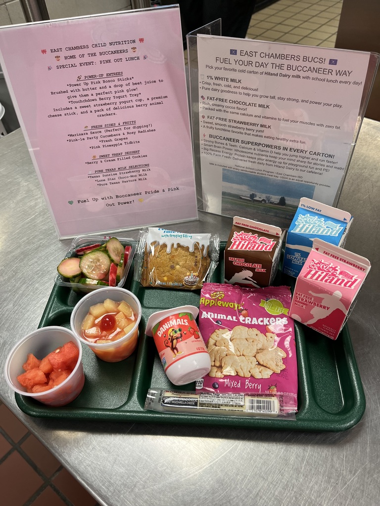 A green tray with various food items: a bowl of watermelon, a cup of juice, a salad, crackers, a yogurt container, and a milk box. A pink sign is behind the tray.