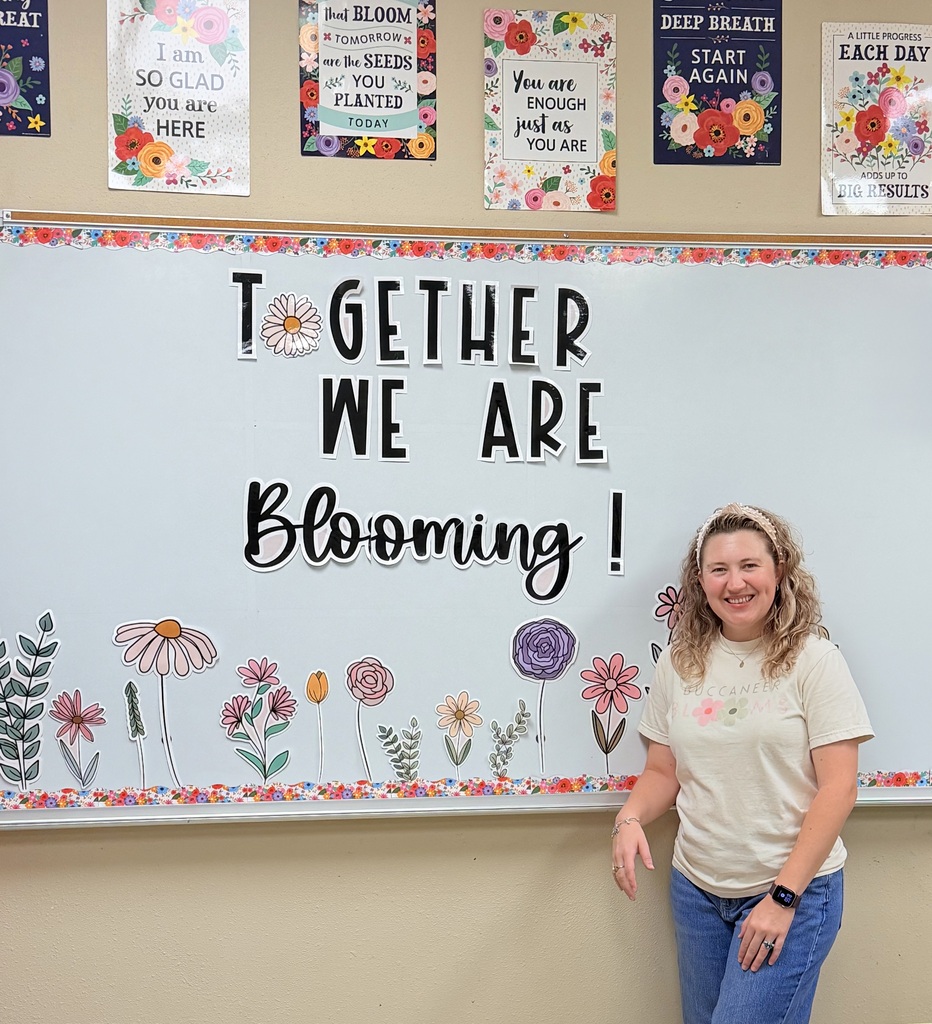 A woman stands before a whiteboard with a floral design. The whiteboard reads "Together We Are Blooming!" She wears a white t-shirt and blue jeans.