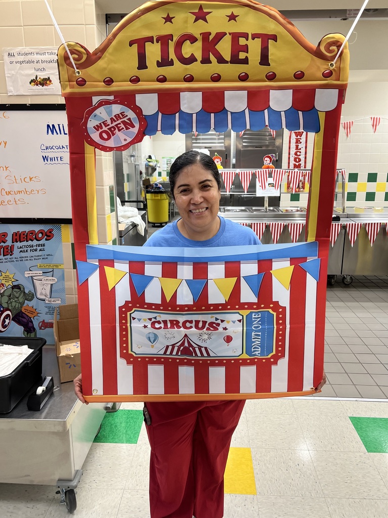 A person stands behind a circus ticket booth, smiling, with a large sign reading "TICKET" on top.