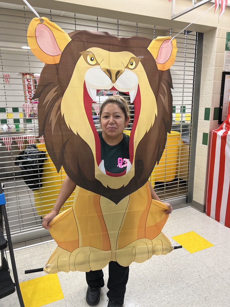 A person wearing a lion costume, including a large head, stands in a hallway. Behind them is a metal gate and a partially visible yellow and red striped curtain.