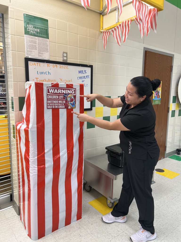 A woman in black clothing and sneakers points to a large red-and-white striped box. Behind her is a bulletin board with text and a door.