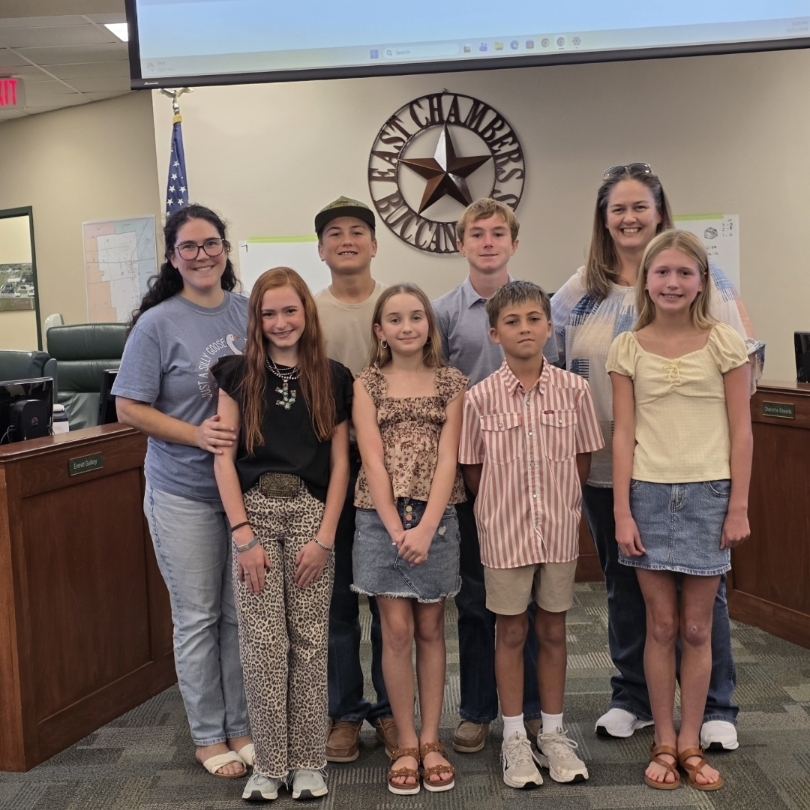 A group of children and adults in a room, possibly a courtroom, with flags and a podium.