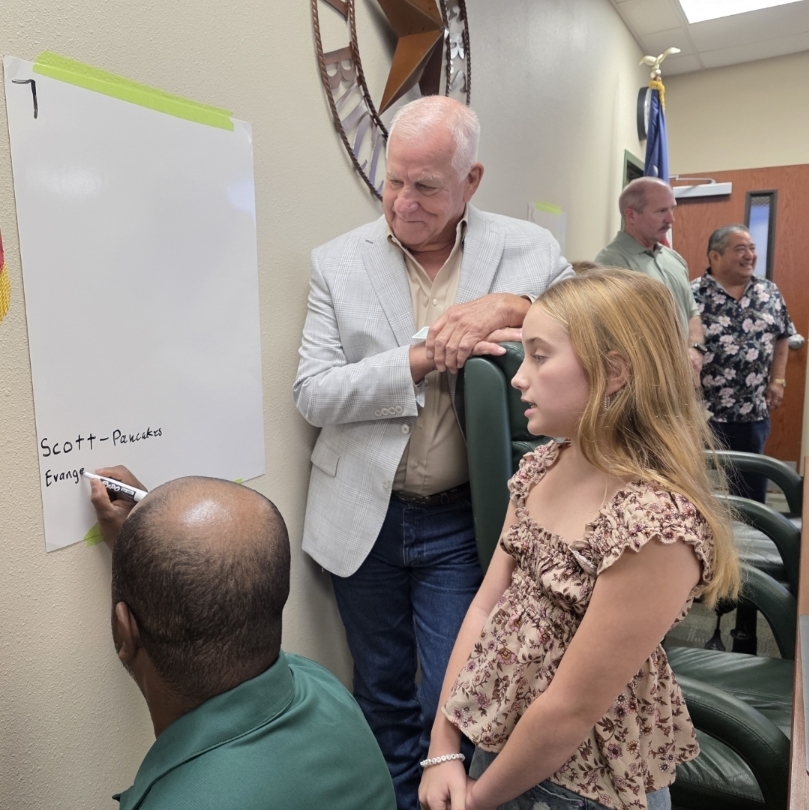 A man signing a whiteboard, with a young girl and another man nearby, an American flag on the left.