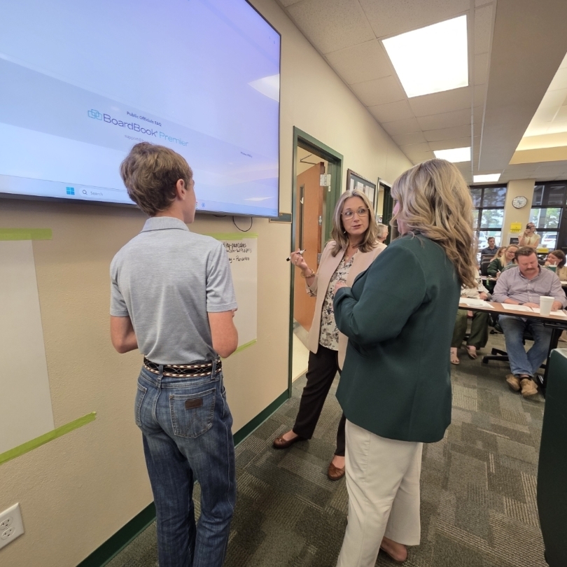 Three people in a corridor. One holds a tablet, another shows a paper, and the third listens.