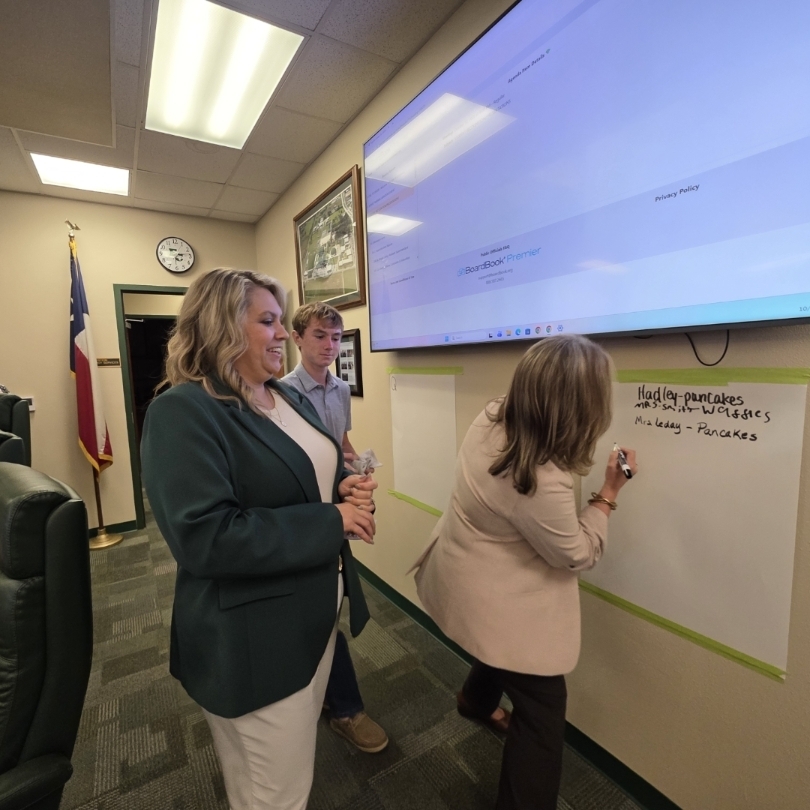 People in a conference room. One woman writes on a whiteboard, another woman watches, and a man stands nearby.