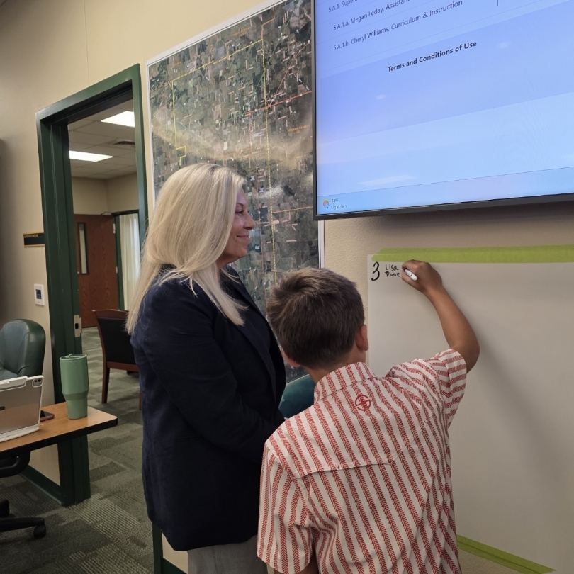 An adult and a child stand by a whiteboard, the child writing. A screen displays behind them.