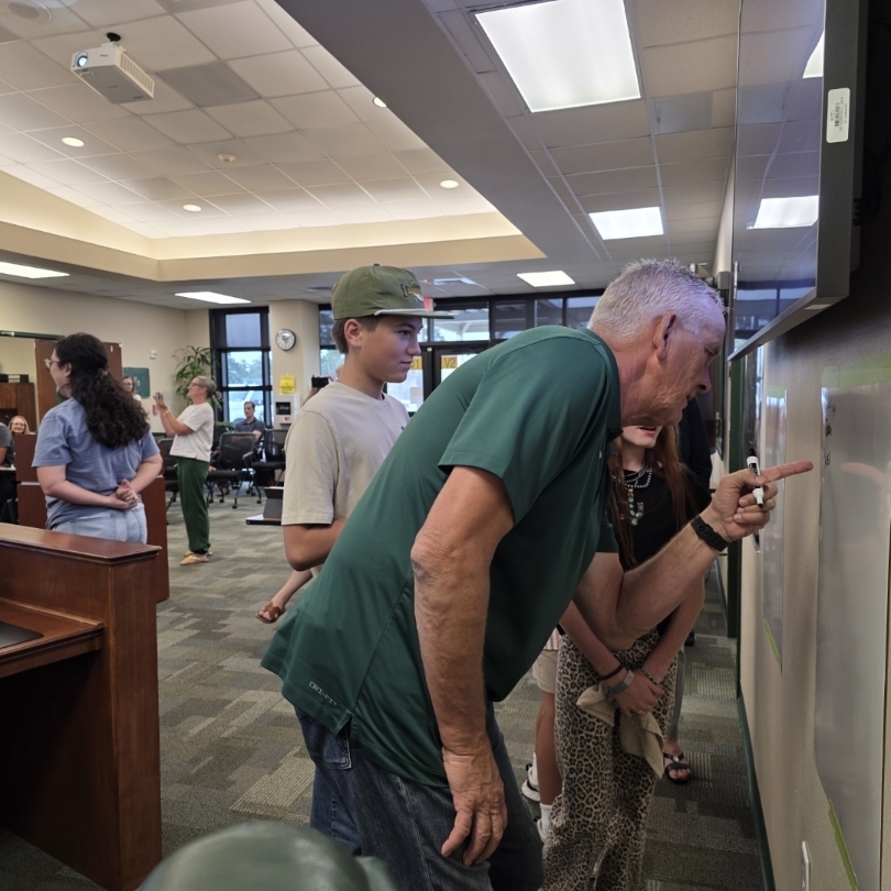 Several people are standing in a room. A man in a green shirt points at a whiteboard on the wall.