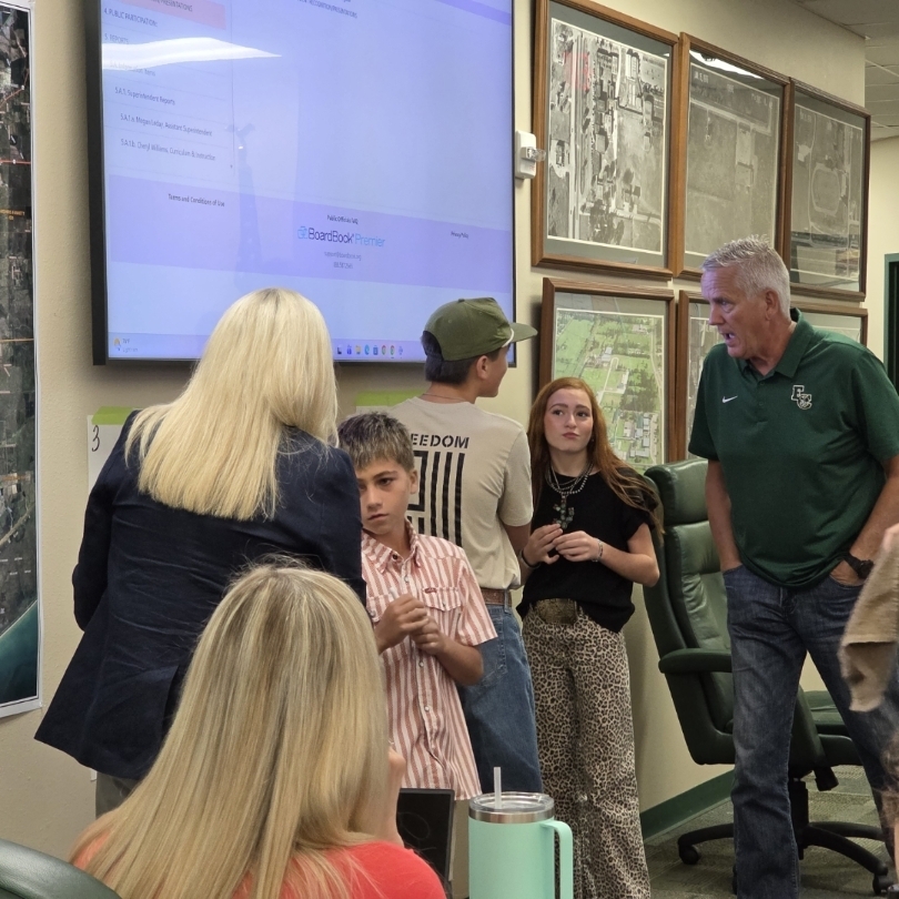 People gather in a room with a large monitor showing a map, maps on the wall, and a man in green.