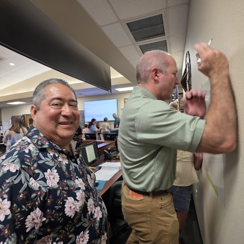 Two men in a room, one standing and writing on a wall, the other smiling and watching.
