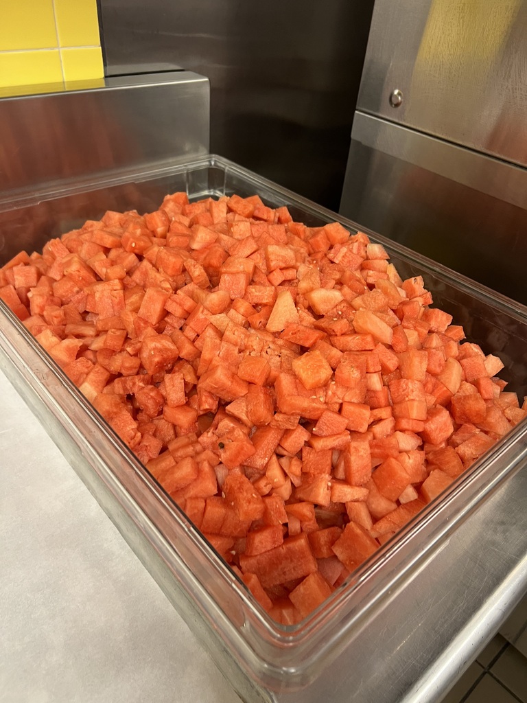 A container filled with cubed watermelons on a metal surface. The container is transparent, and the watermelons are orange.