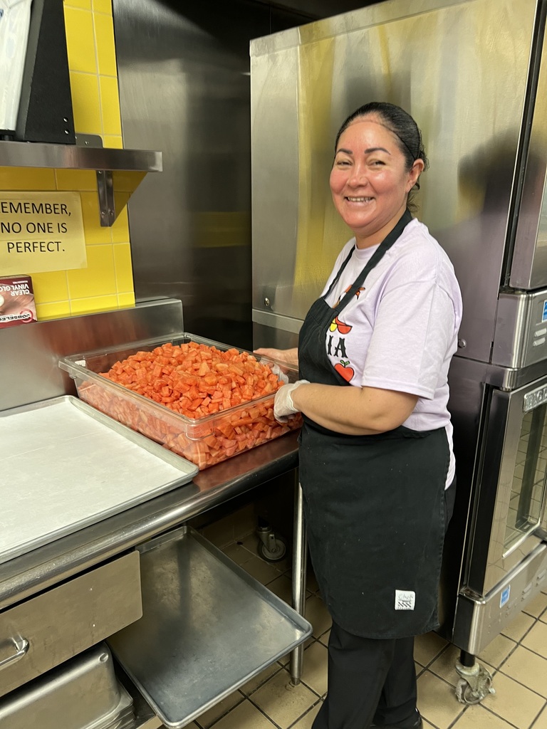 Woman in apron stands in kitchen, holding a tray of red food, next to refrigerators.