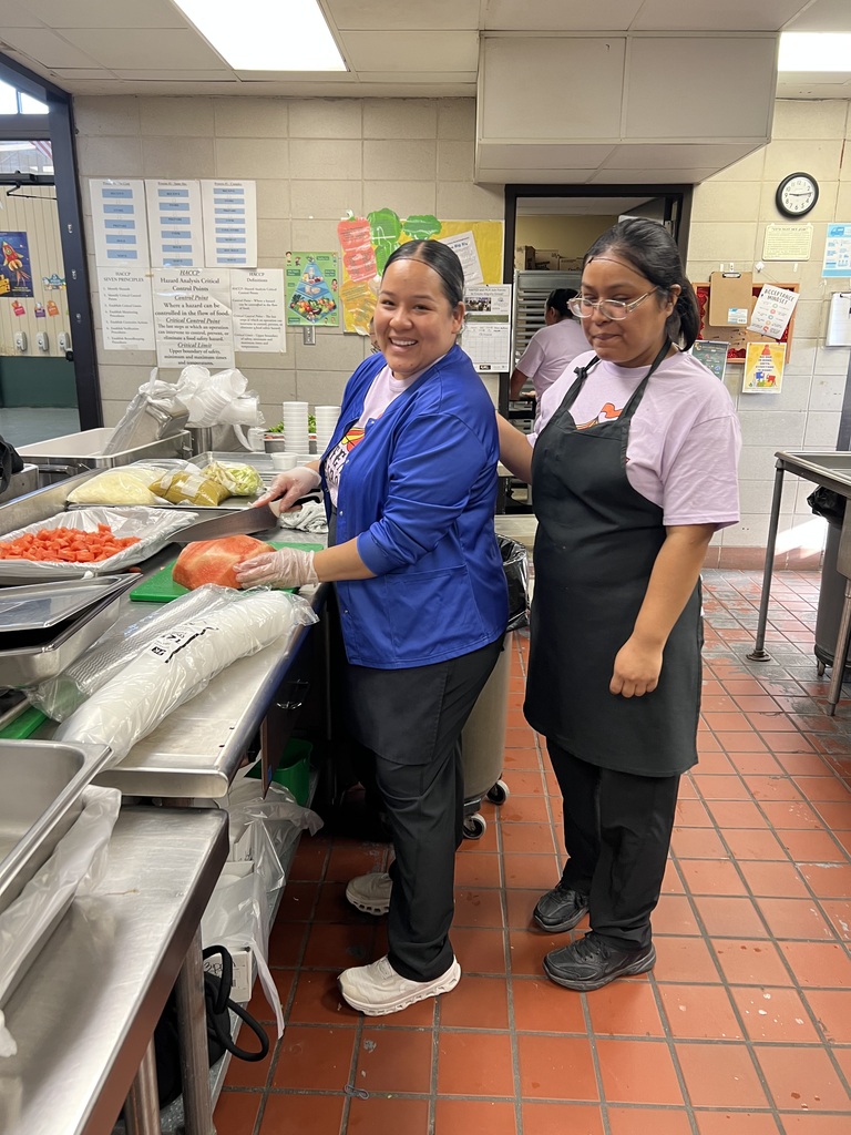 Two women in a kitchen. One holds a knife and the other wears glasses. Trays and bowls are on the counter.