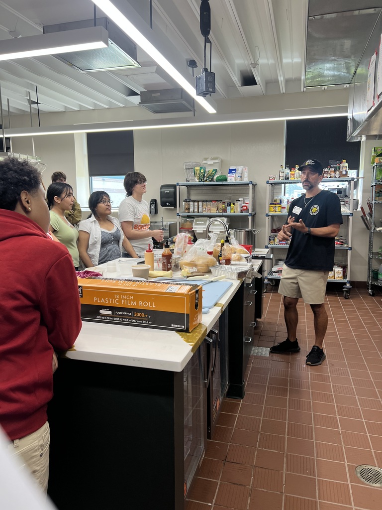 Group of people in a kitchen. One person speaks while others listen. Counter has food items.