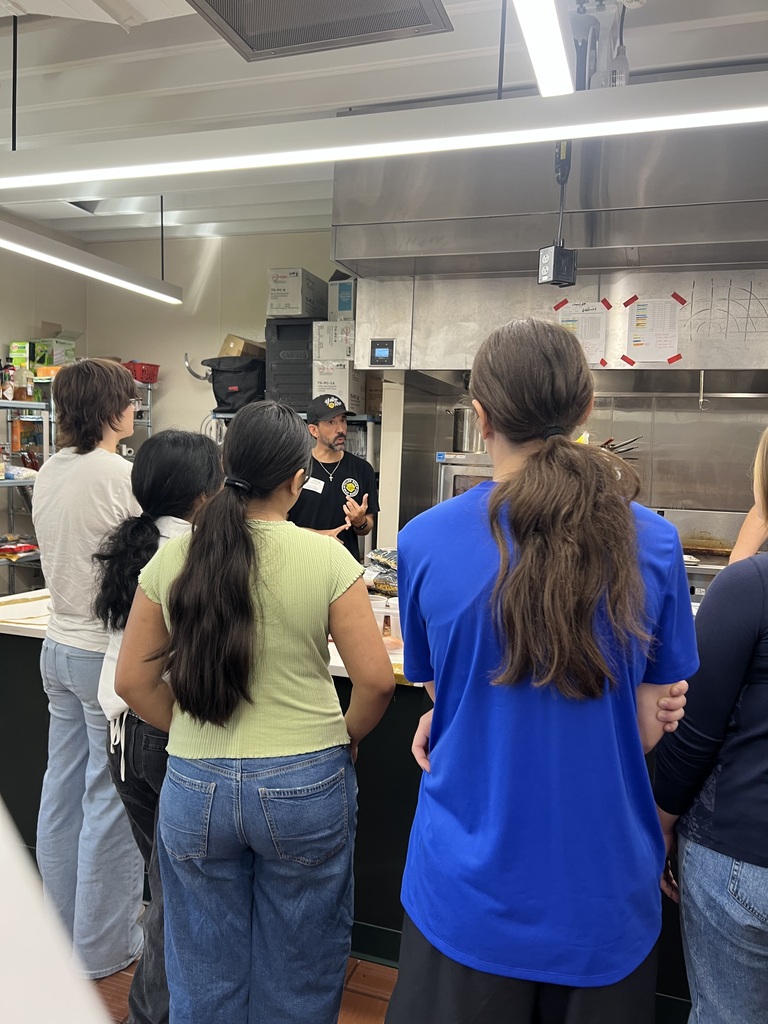 People stand in a kitchen, listening to a person speaking. The kitchen has a countertop, shelves, and overhead lights.