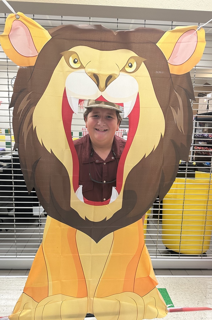 A person stands behind a lion cutout, smiling, with a yellow trash bin behind them and a metal grid in the background.