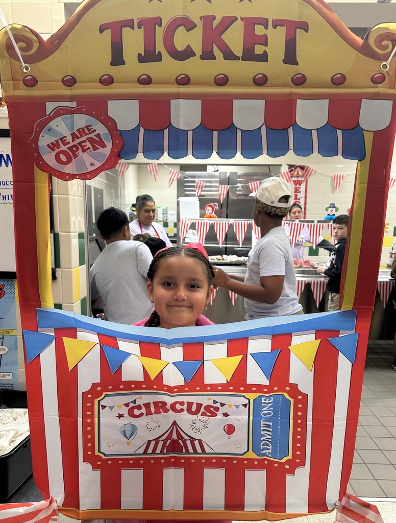 A girl is standing behind a colorful booth with a ticket window and a circus theme. People are in the background.