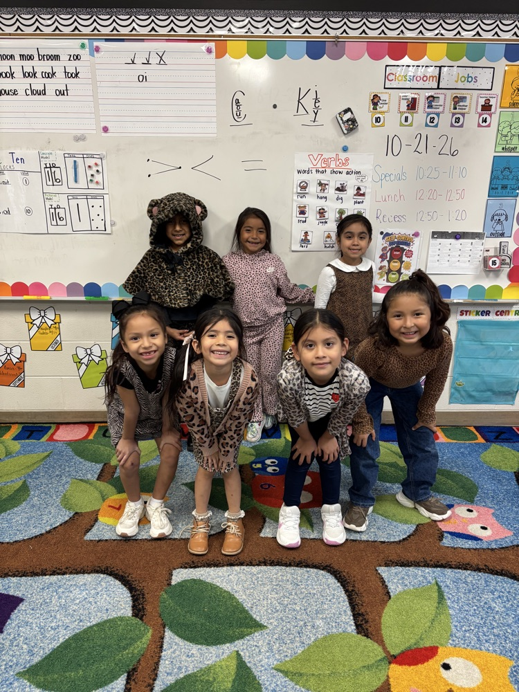 Six young girls pose for a picture with one in a bear costume in a classroom.