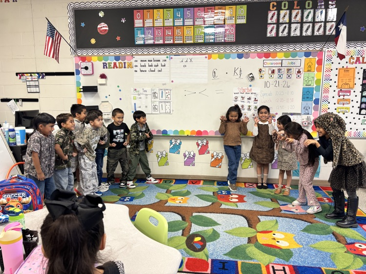Young children stand in a circle in front of a teacher; they appear to be listening.