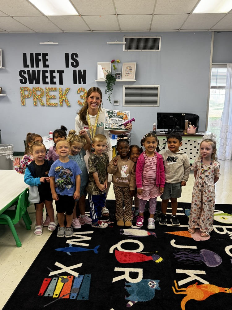 Preschool class with children standing on a rug that says 'My World'. A teacher holds a book.