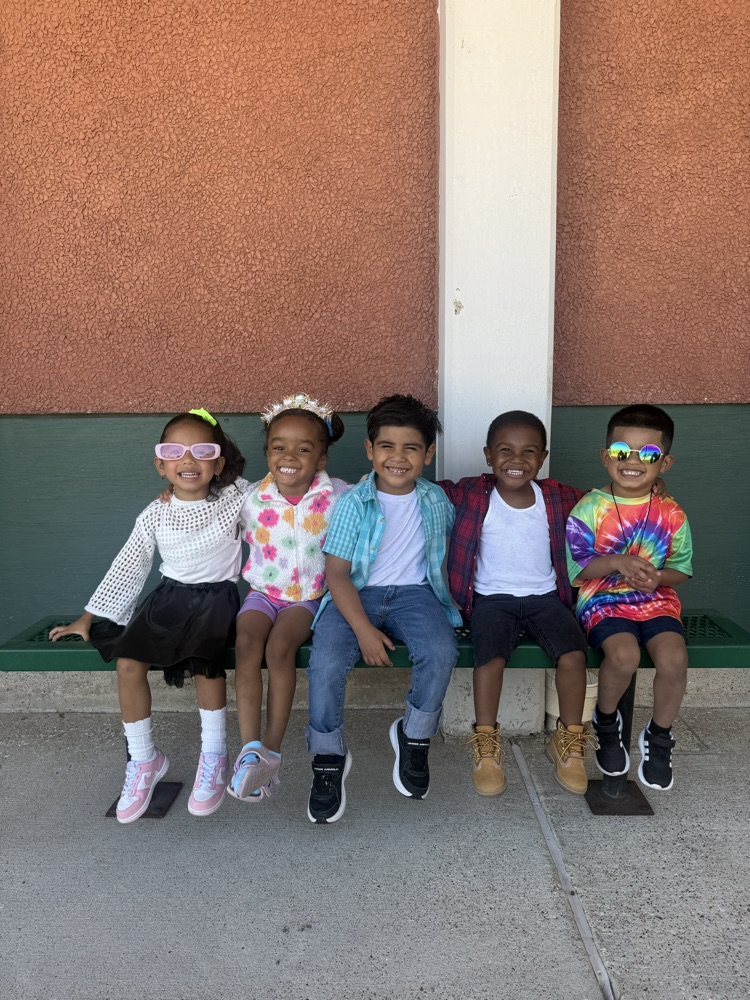 Five children in different colored shirts sit on a bench against a red wall. They are wearing sunglasses and various sneakers.