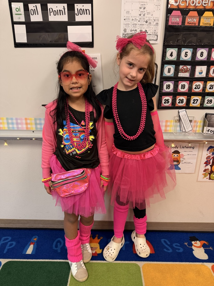 Two young girls in pink tutus, socks, and glasses stand in front of a bulletin board.